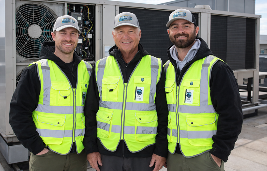 three men wearing yellow vests in front of an air handler